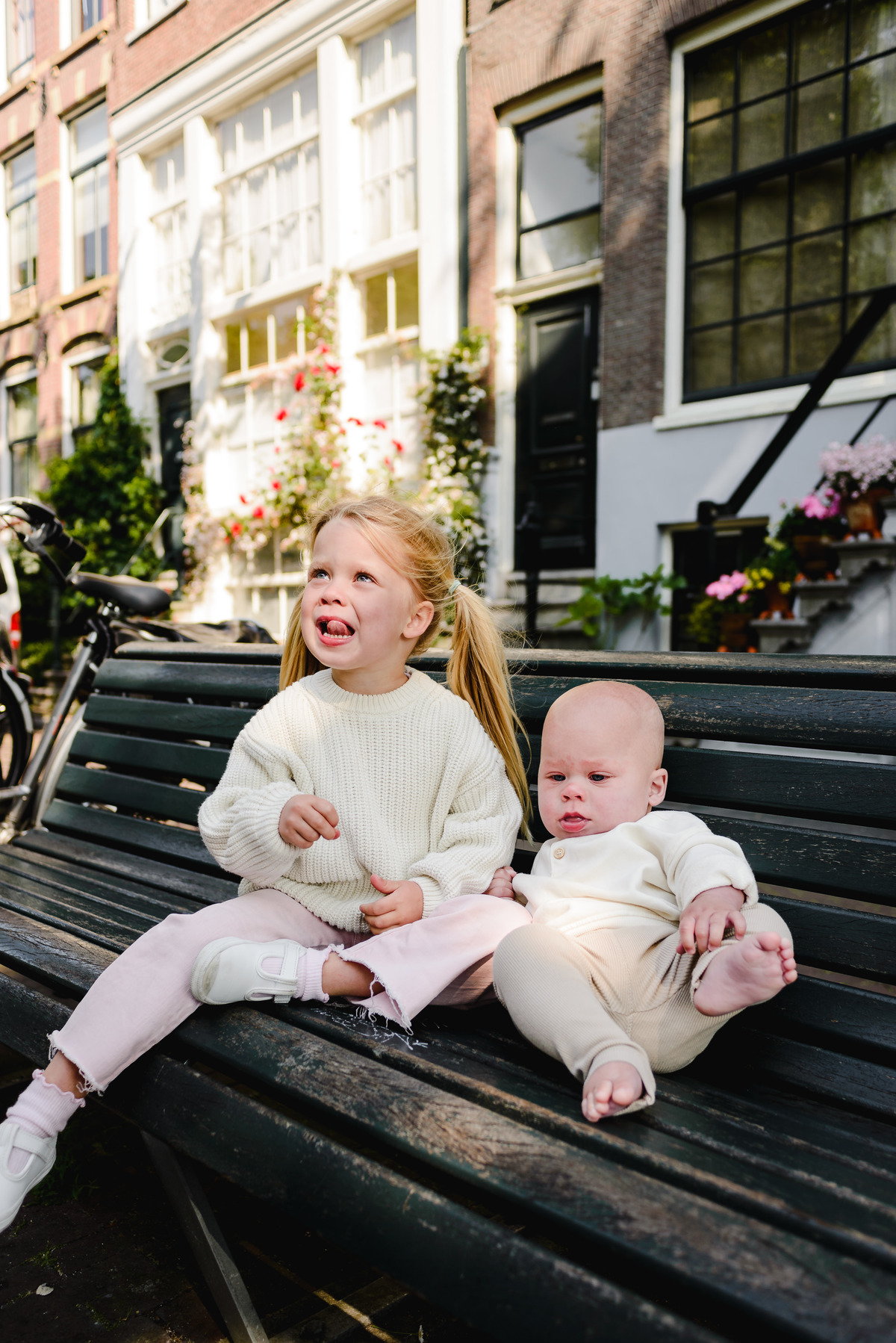 Two young siblings sitting on a bench along an Amsterdam canal, with historic canal houses and greenery in the background on a sunny day.