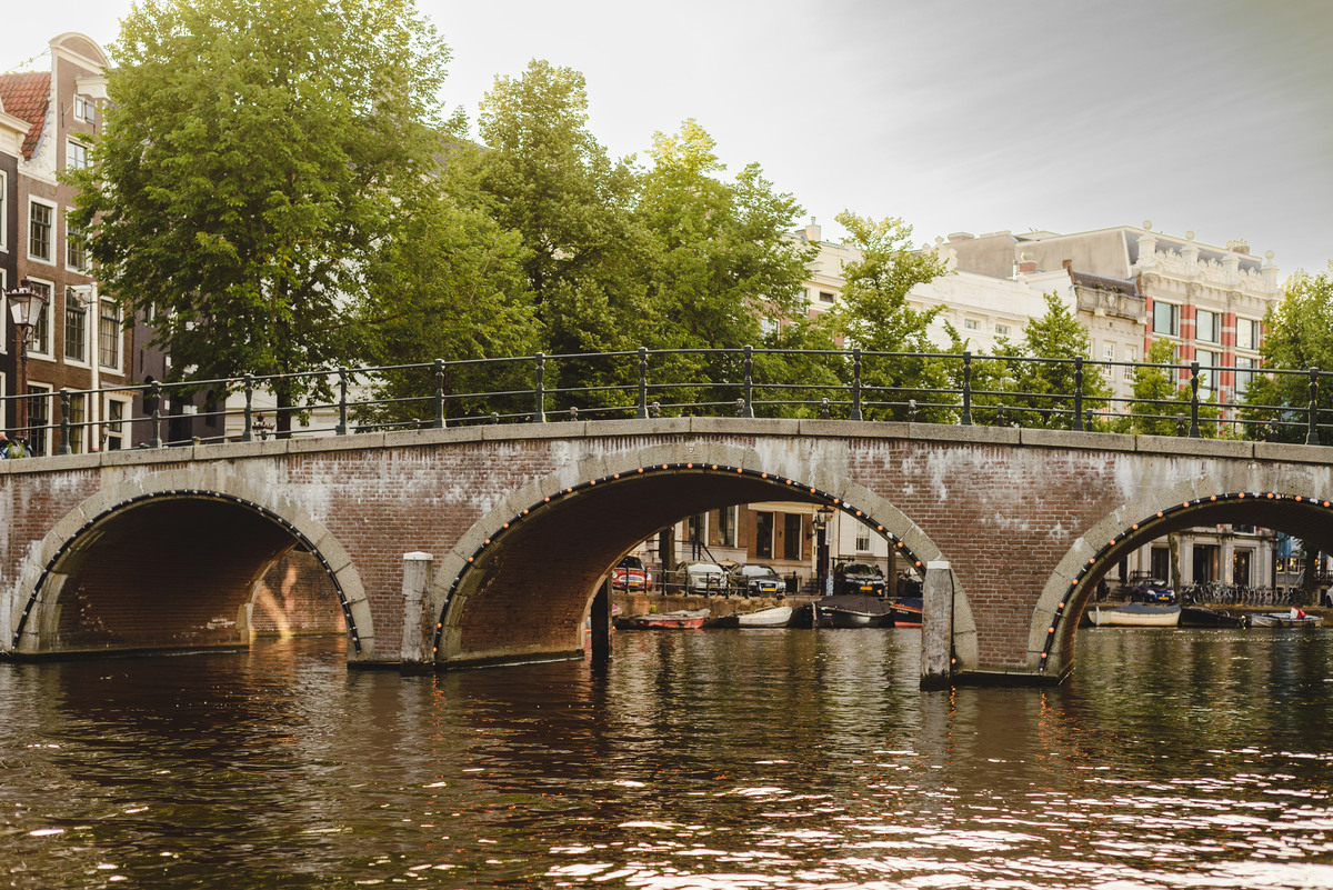 Wide view of an Amsterdam canal with a brick bridge, houseboats, and leafy streets in summer.