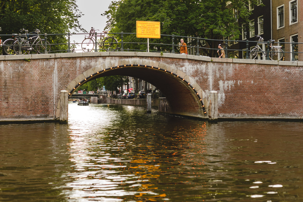 Brick arch bridge over an Amsterdam canal with trees and historic houses in summer light.