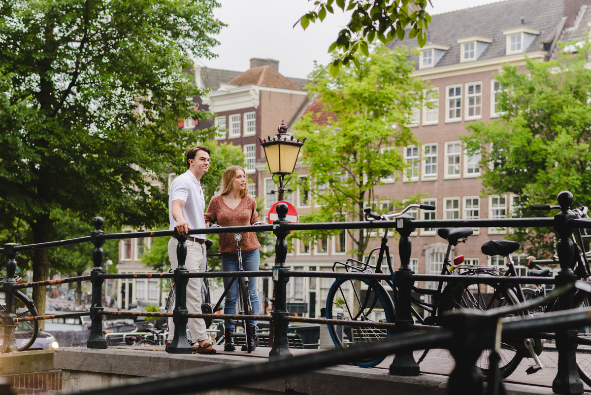 Couple standing on a small Amsterdam bridge with canal and houseboats behind them on a cloudy summer day.