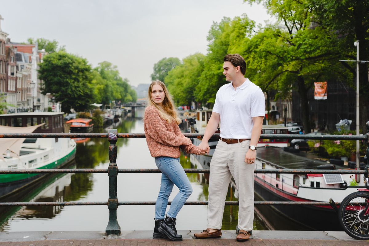 Couple standing on a small Amsterdam bridge with canal and houseboats behind them on a cloudy summer day.
