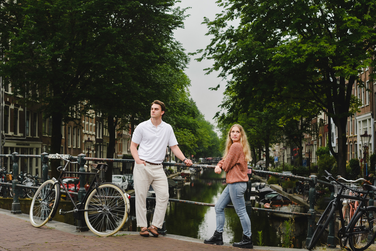 Couple standing on a small Amsterdam bridge with canal and houseboats behind them on a cloudy summer day.