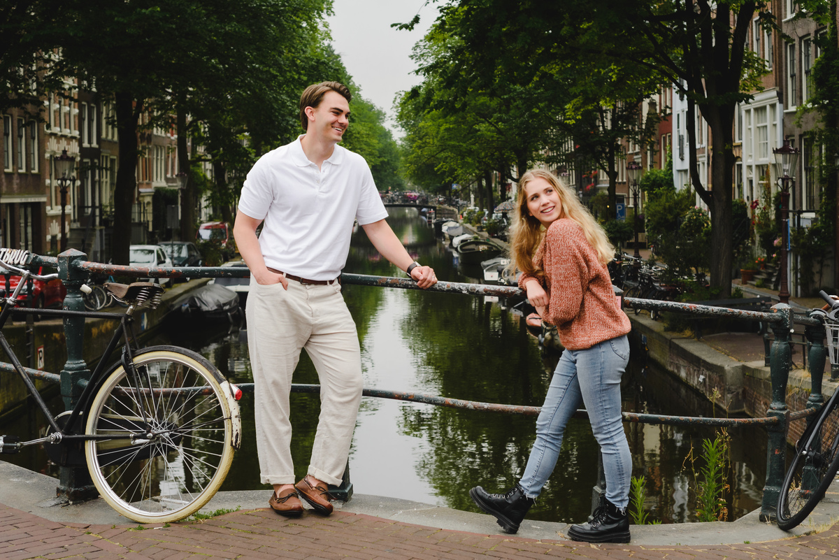 Couple standing on a small Amsterdam bridge with canal and houseboats behind them on a cloudy summer day.