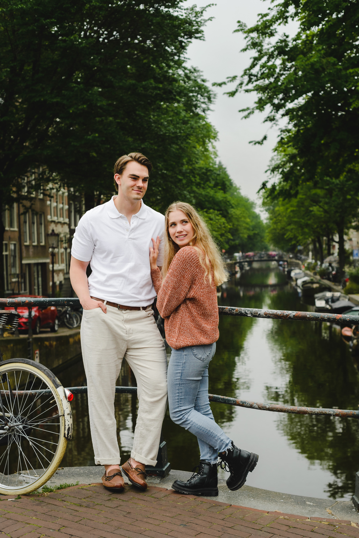 Couple standing on a small Amsterdam bridge with canal and houseboats behind them on a cloudy summer day.