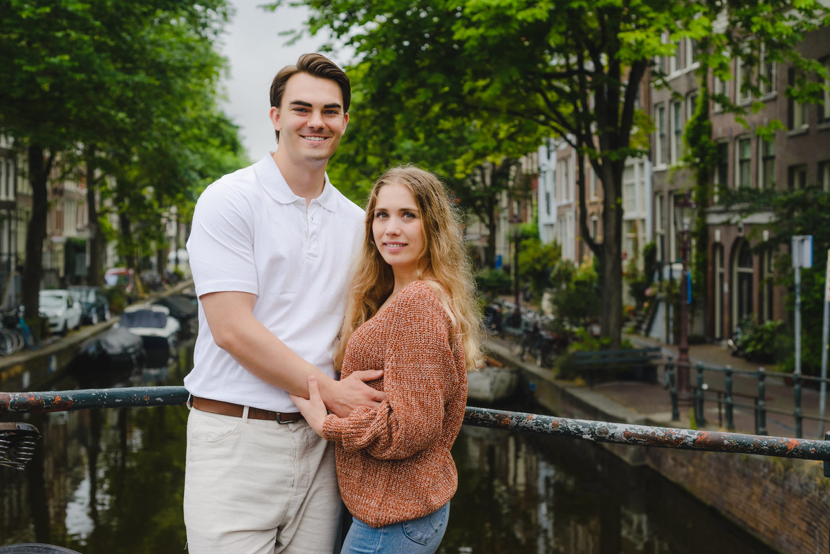 Couple standing on a small Amsterdam bridge with canal and houseboats behind them on a cloudy summer day.