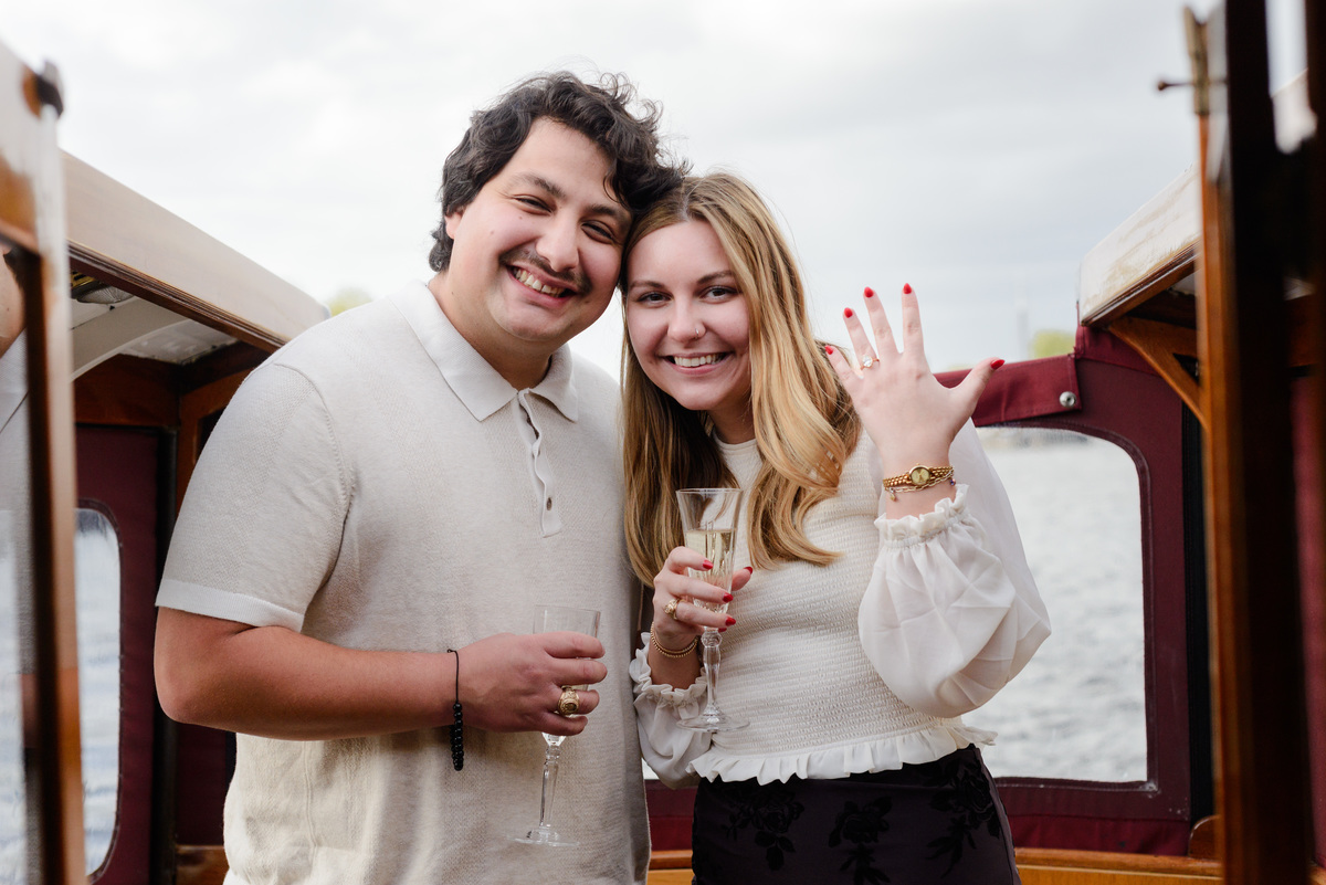 A woman is showing a serene smile to a camera next to the fiancé. They are recently engaged and toasting while navigating the canals of Amsterdam. 