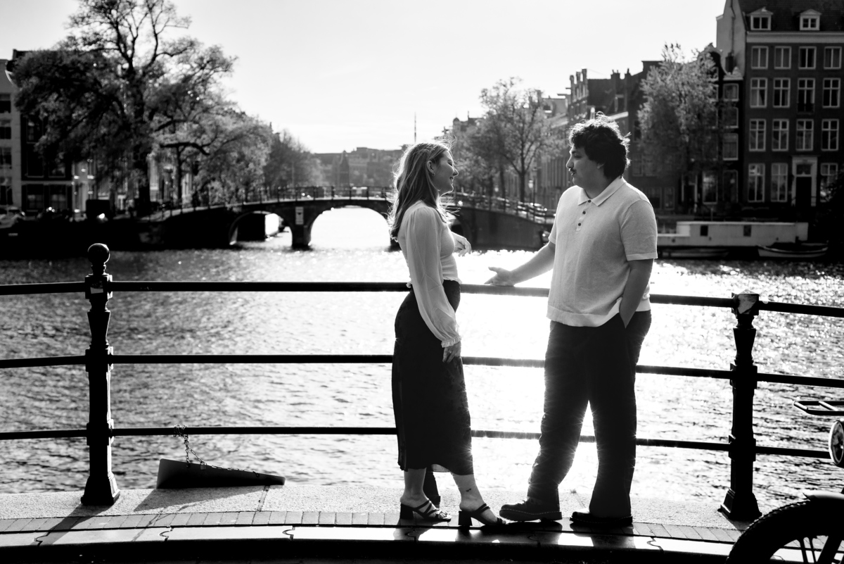 A black and white image backlighted of the couple standing on a bridge in Amsterdam. You see the Amstel river behind them. 