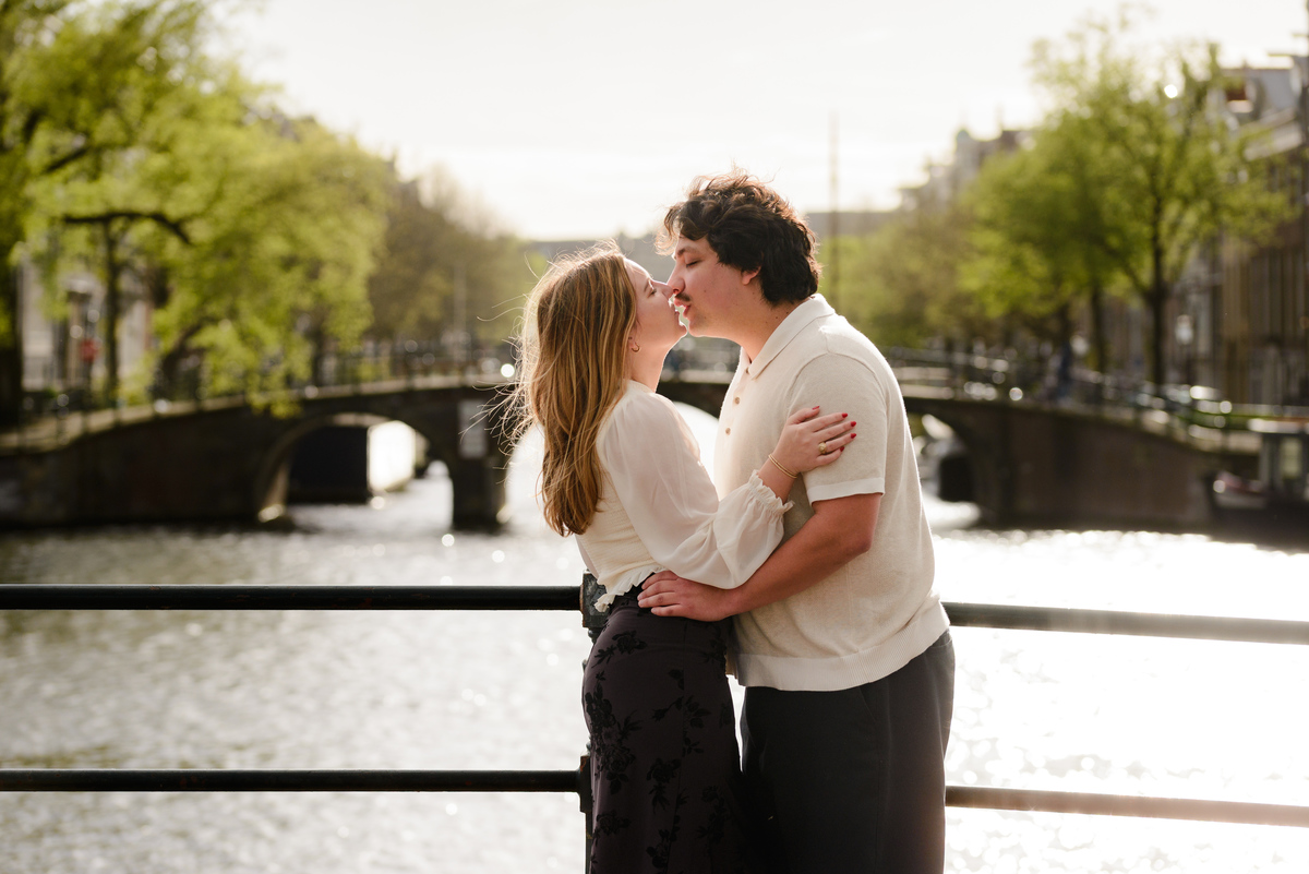 A couple share a kiss with the Amstel river behind them. You can see a bridge, an Amsterdam canal bridge, behind them. 