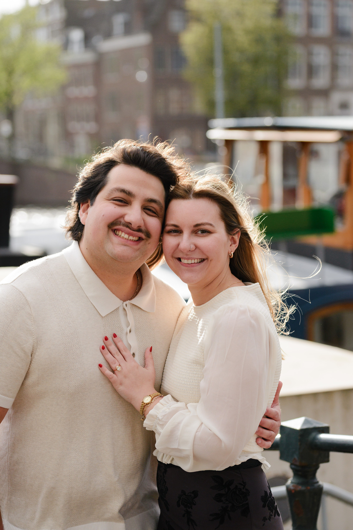 A happy couple smiling to the camera with a beautiful Amsterdam background behind them. They are recently engaged. He proposed to her on a canal cruise in Amsterdam. 