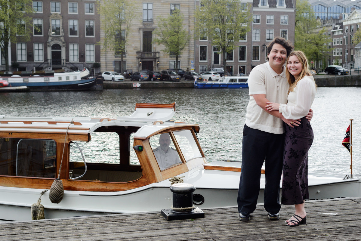A young couple recently engaged in the canals of Amsterdam is posing next to their boat, which is a historical ship from 1932. 