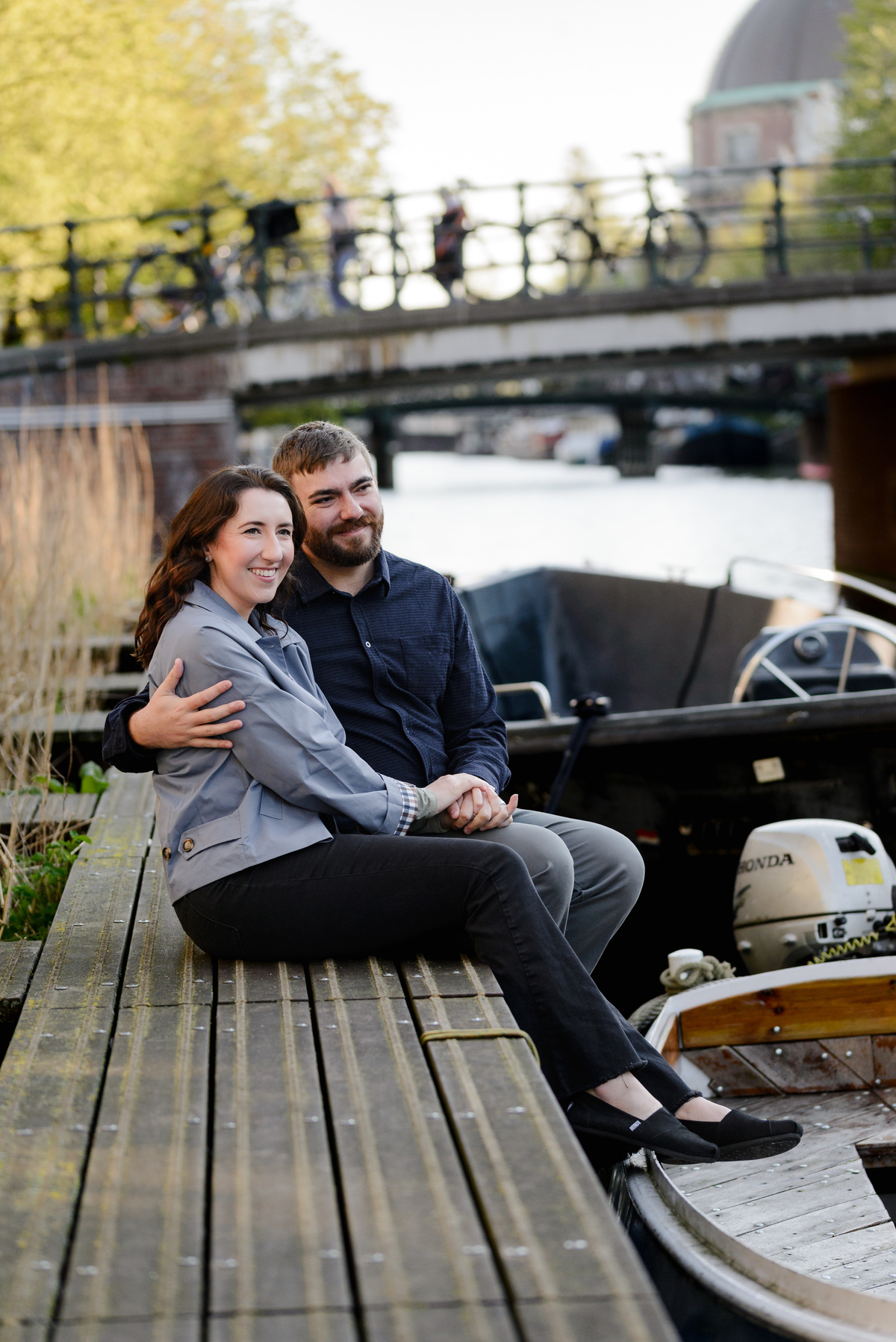 Happy couple sitting next to a canal in Amsterdam with boats in the background and a bridge with bikes 