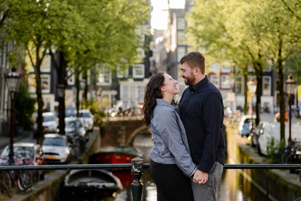 Couple holding hands with the background of the canal bell of Amsterdam, a beautiful bridge with bikes and visible boats behind them. This is a photo session in April 2026. 