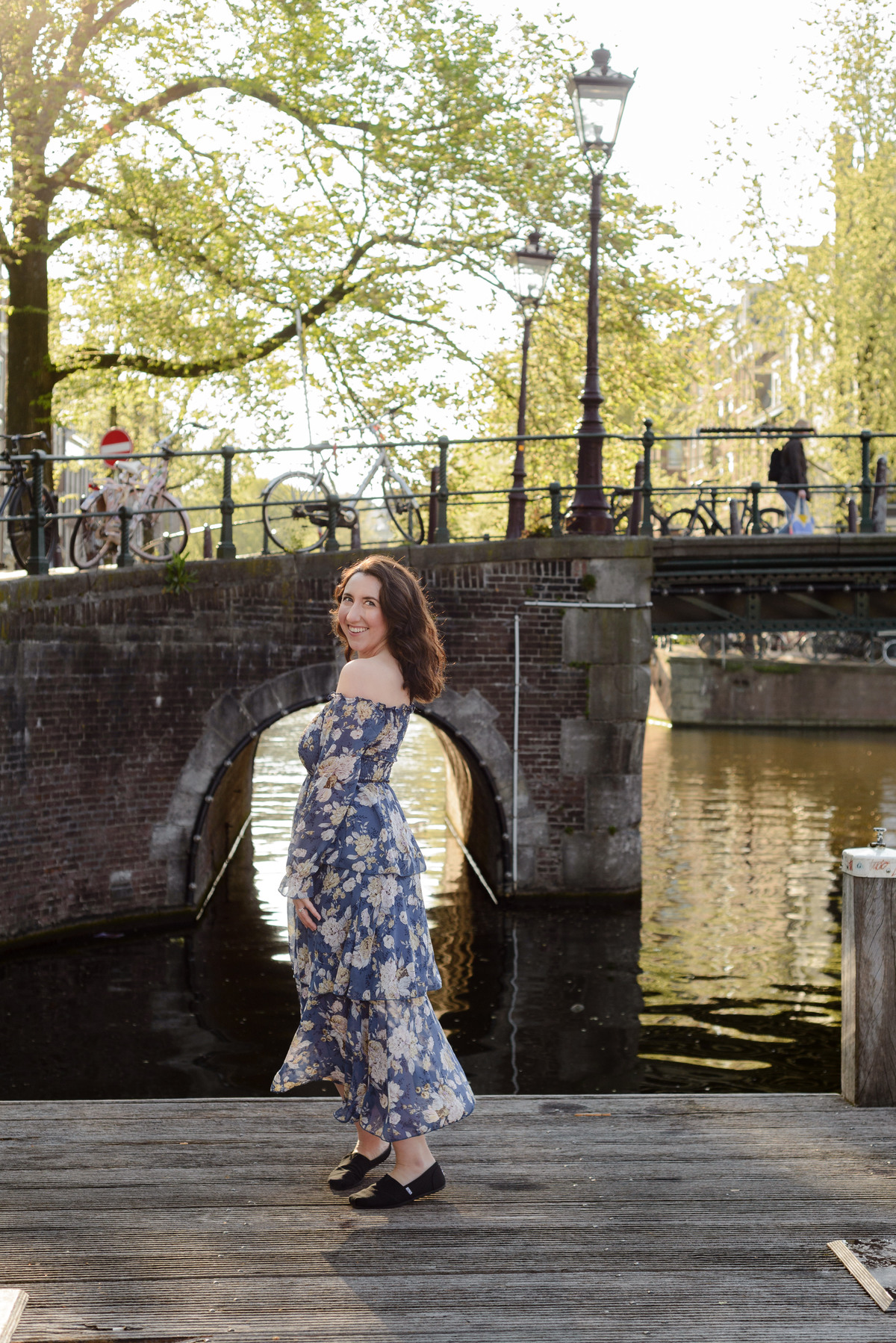 A woman smiling at the camera with a beautiful floral dress. The background is a historical canal in Amsterdam with bikes. This was taken during a photo session in Amsterdam, in one of the best locations to photograph in Amsterdam. 
