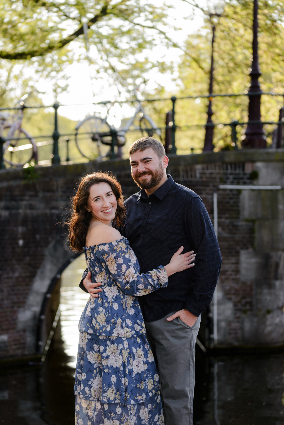 Happy couple smiles at the camera while hugging during a photo session with a beautiful bridge in the background with bikes. 