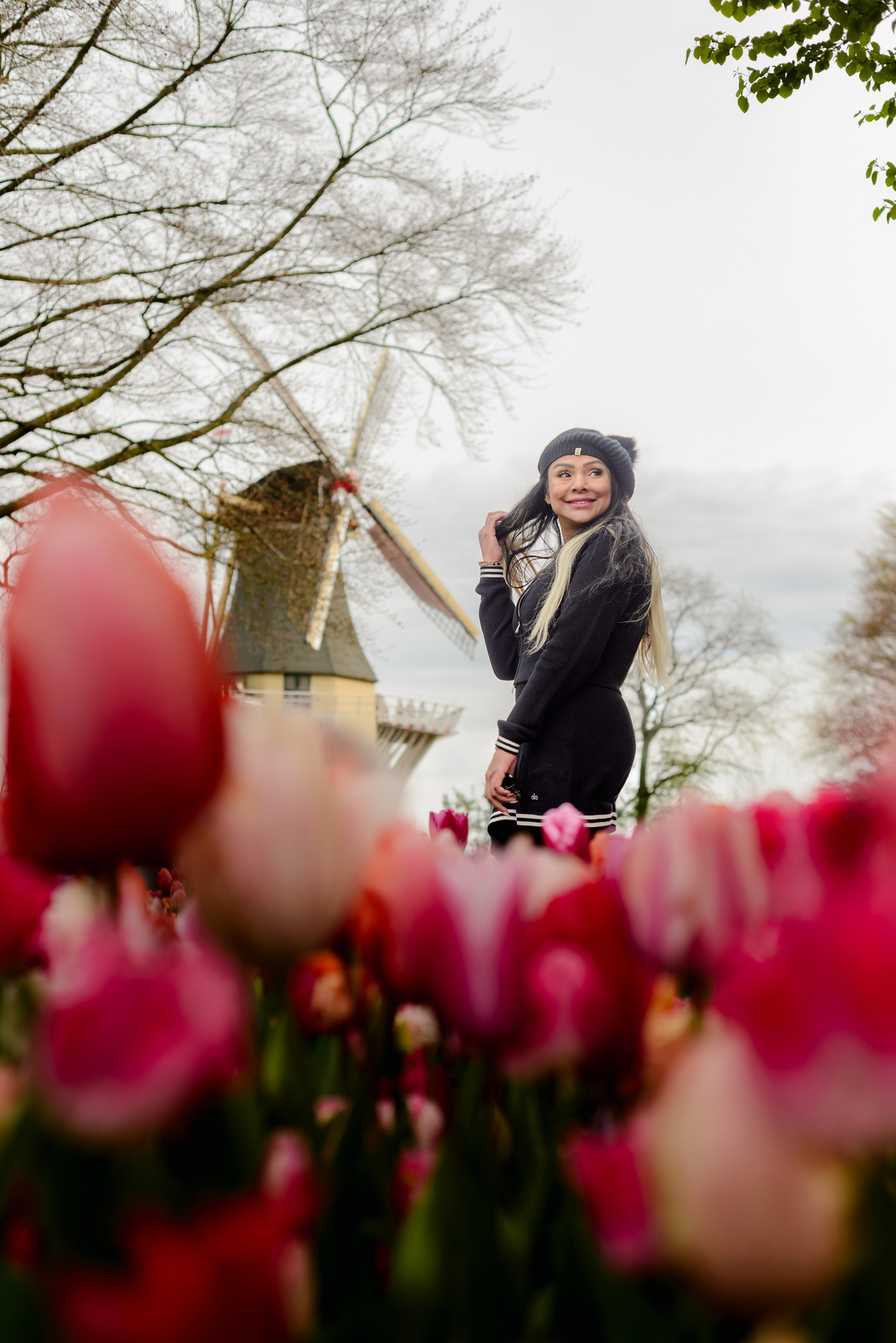 Female solo traveler smiling during a photo session in Keukenhof Gardens, with tulips in front of her and an iconic windmill behind her. 