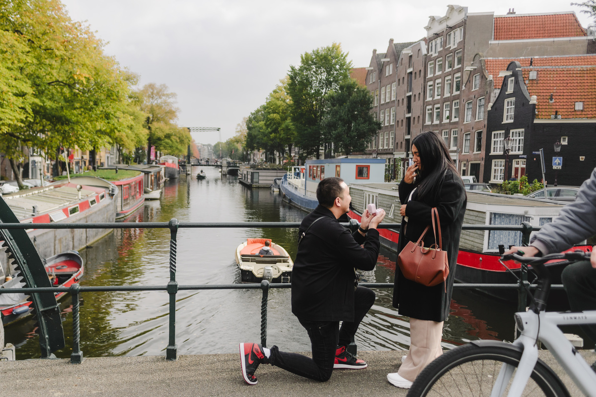 Man kneeling during a surprise marriage proposal on Dommersbrug bridge over Brouwersgracht in Amsterdam while his partner reacts with surprise.