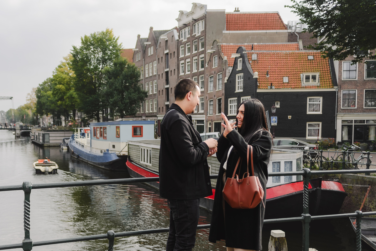 Man kneeling during a surprise marriage proposal on Dommersbrug bridge over Brouwersgracht in Amsterdam while his partner reacts with surprise.
