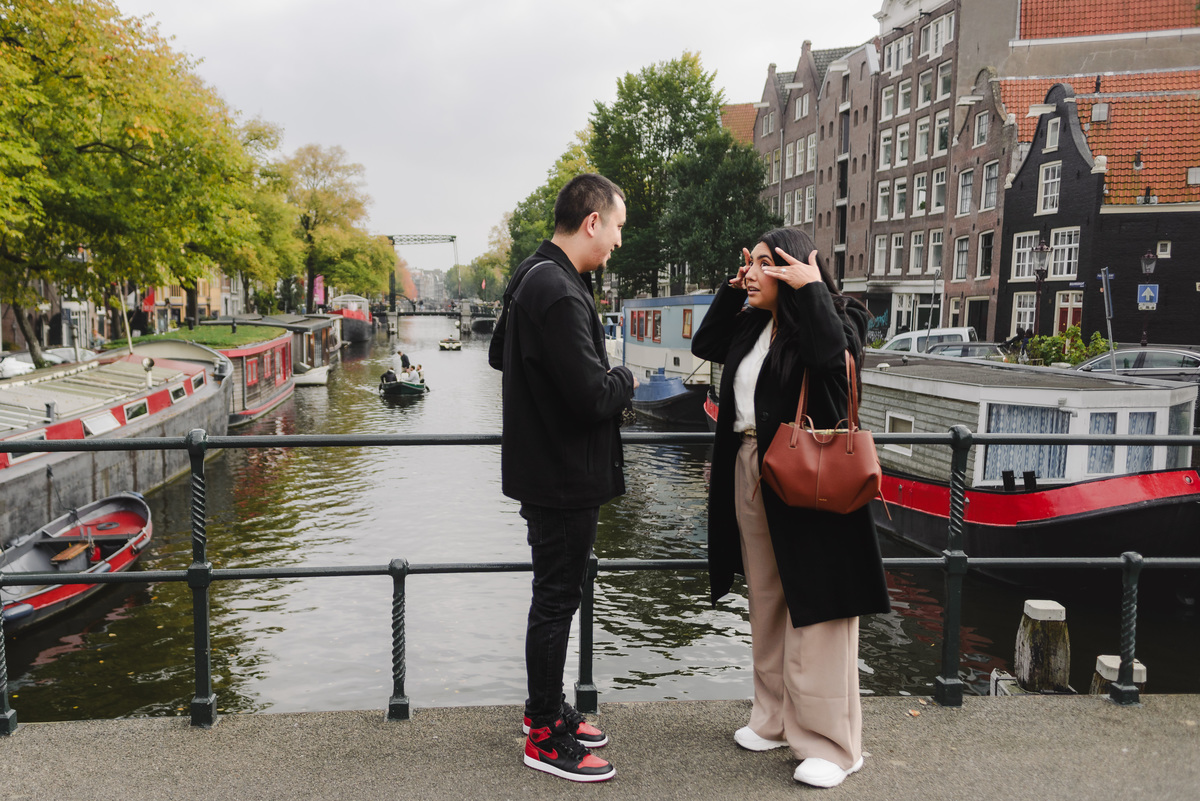 Man kneeling during a surprise marriage proposal on Dommersbrug bridge over Brouwersgracht in Amsterdam while his partner reacts with surprise.