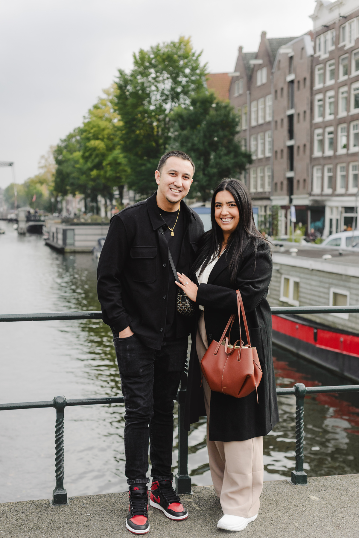 Recently engaged couple smiling together on Dommersbrug with Brouwersgracht canal and historic Amsterdam houses in the background.