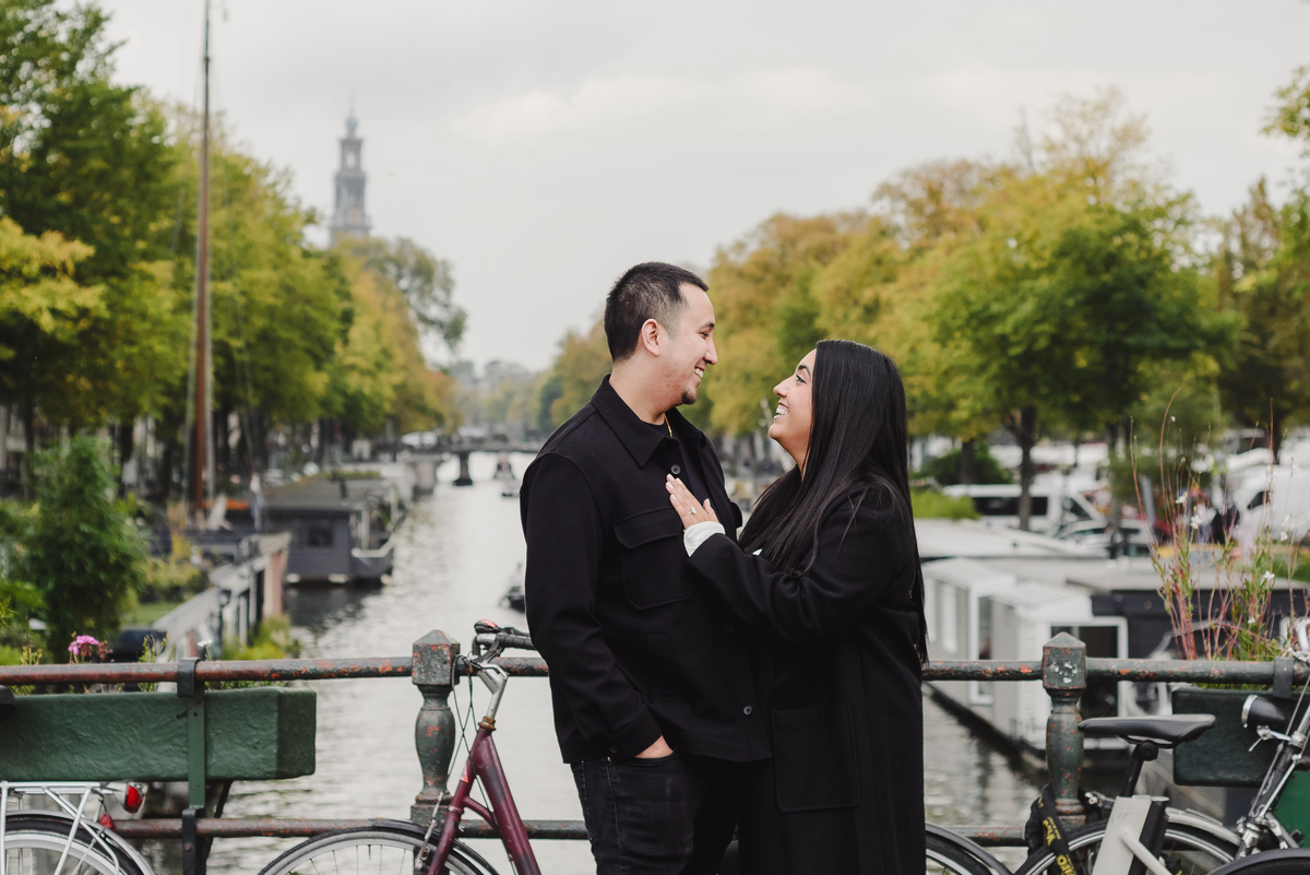 Engaged couple standing on a bridge over Prinsengracht with bicycles and historic canal houses in the background.