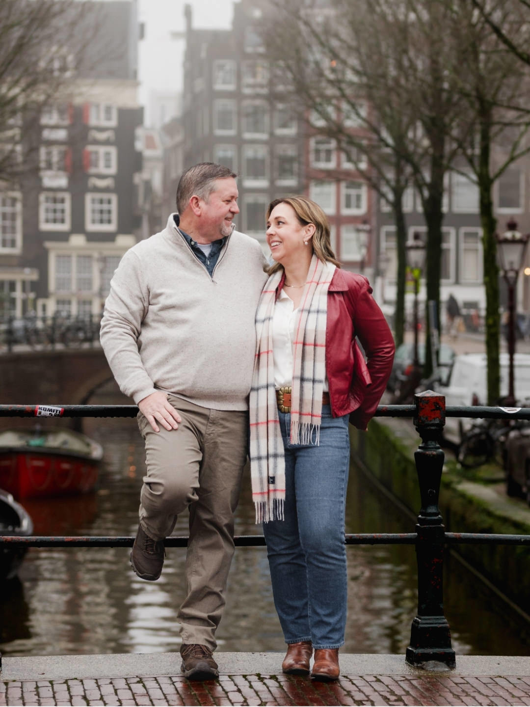 Couple smiling together on a canal bridge with traditional Amsterdam architecture in the background.
