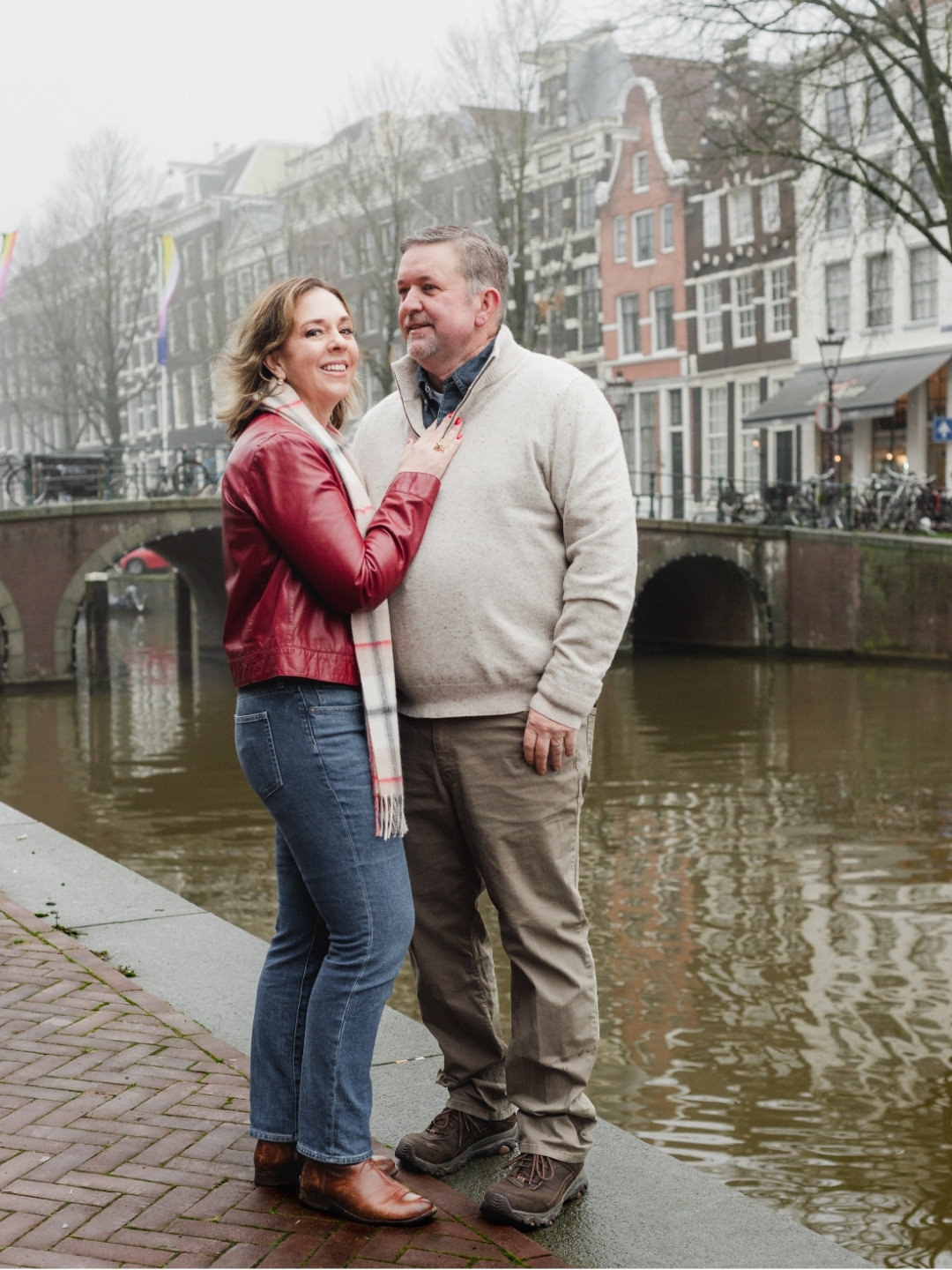 Couple embracing beside an Amsterdam canal with historic bridges and canal houses in the background.