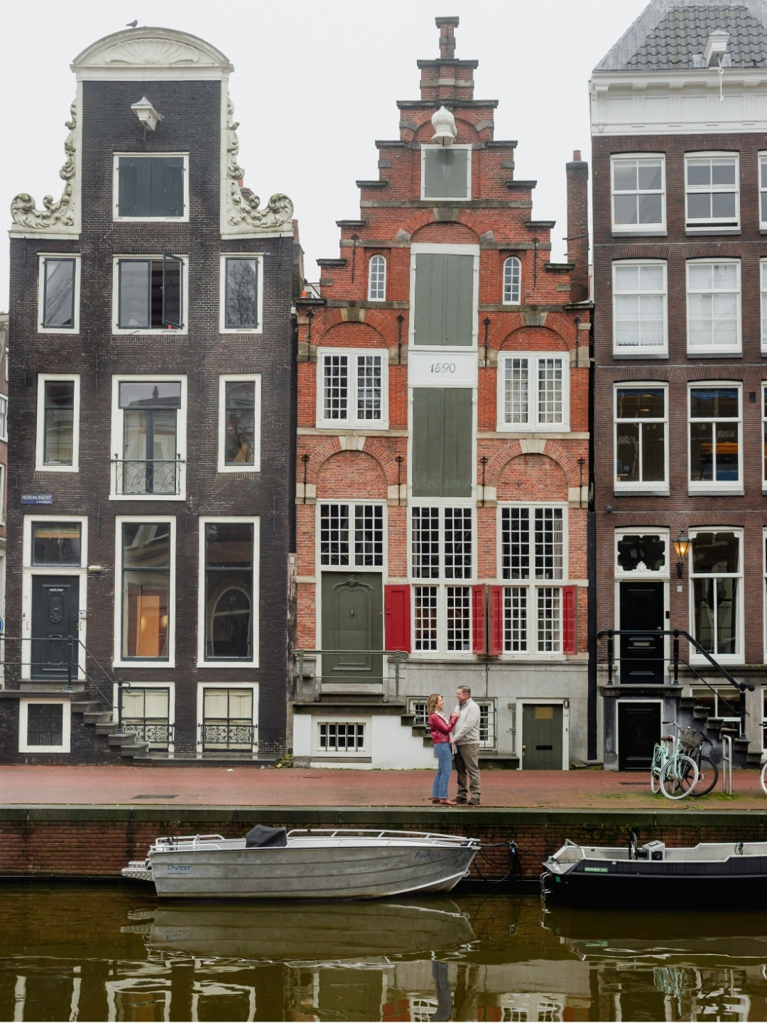 Couple embracing beside an Amsterdam canal with historic bridges and canal houses in the background.