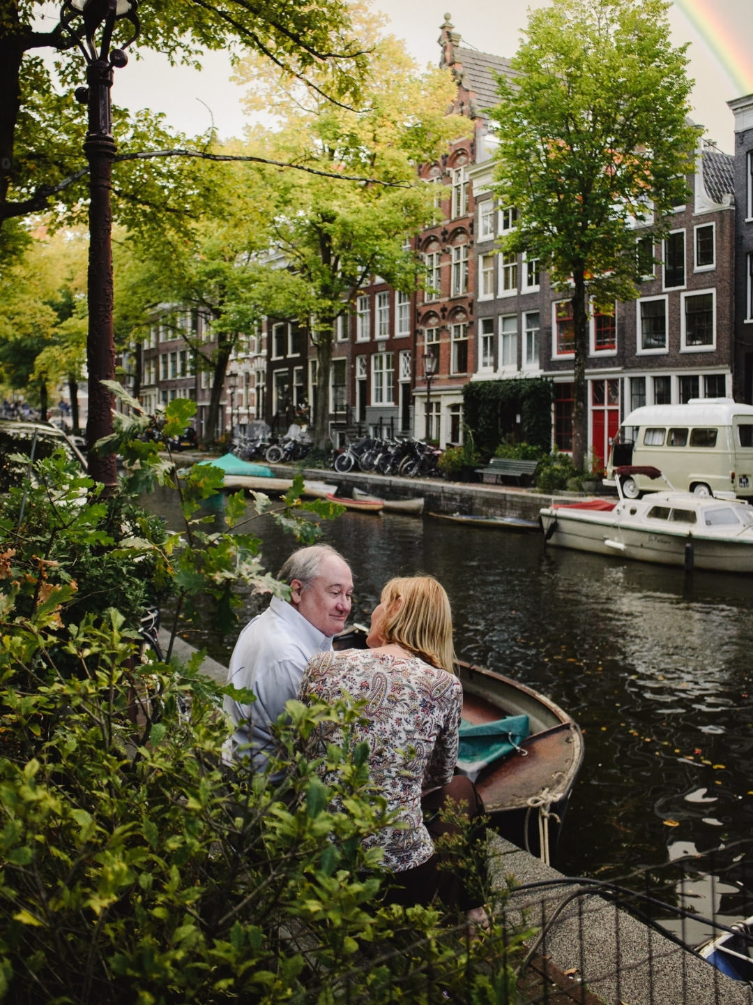 Couple sitting beside a canal in Amsterdam surrounded by greenery and historic architecture.
