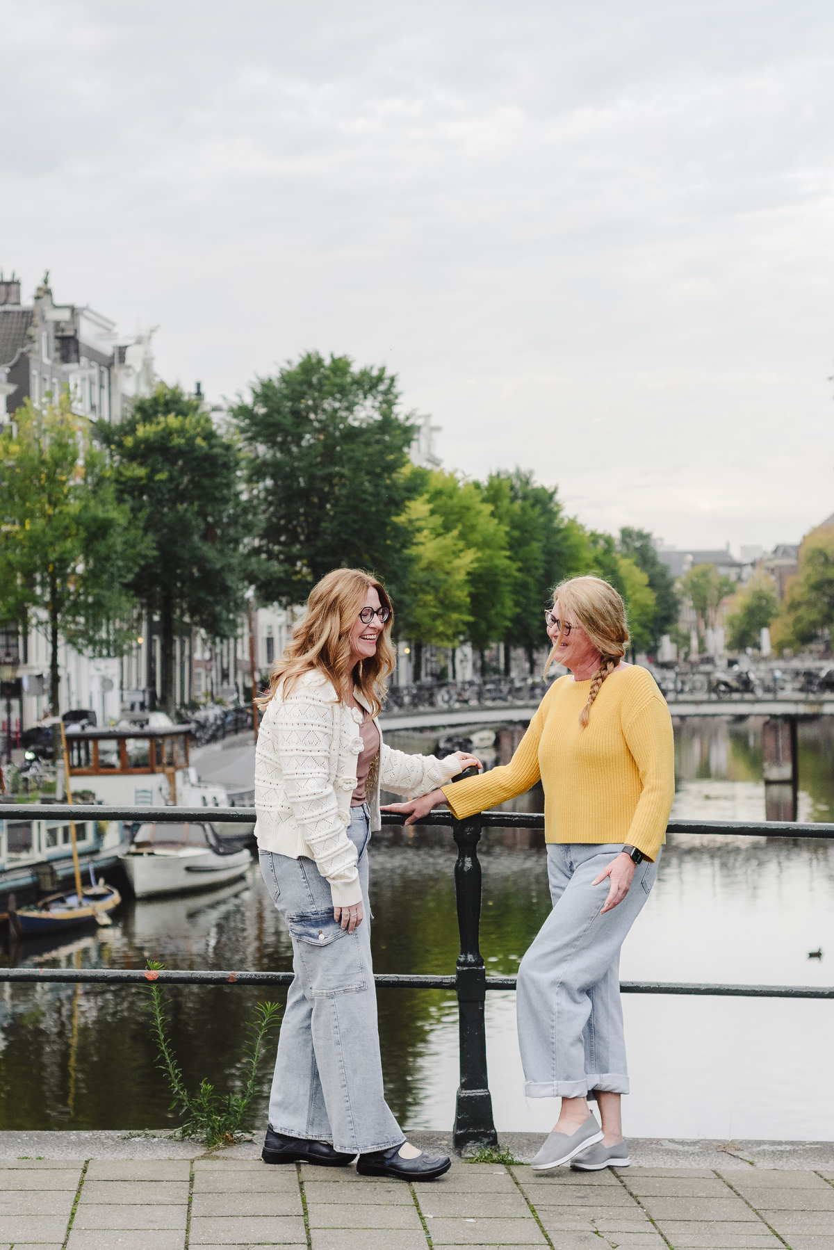Two sisters smiling to each other in front of a canal in Amsterdam with historical houses and bikes in the background. This was a morning photo session. 