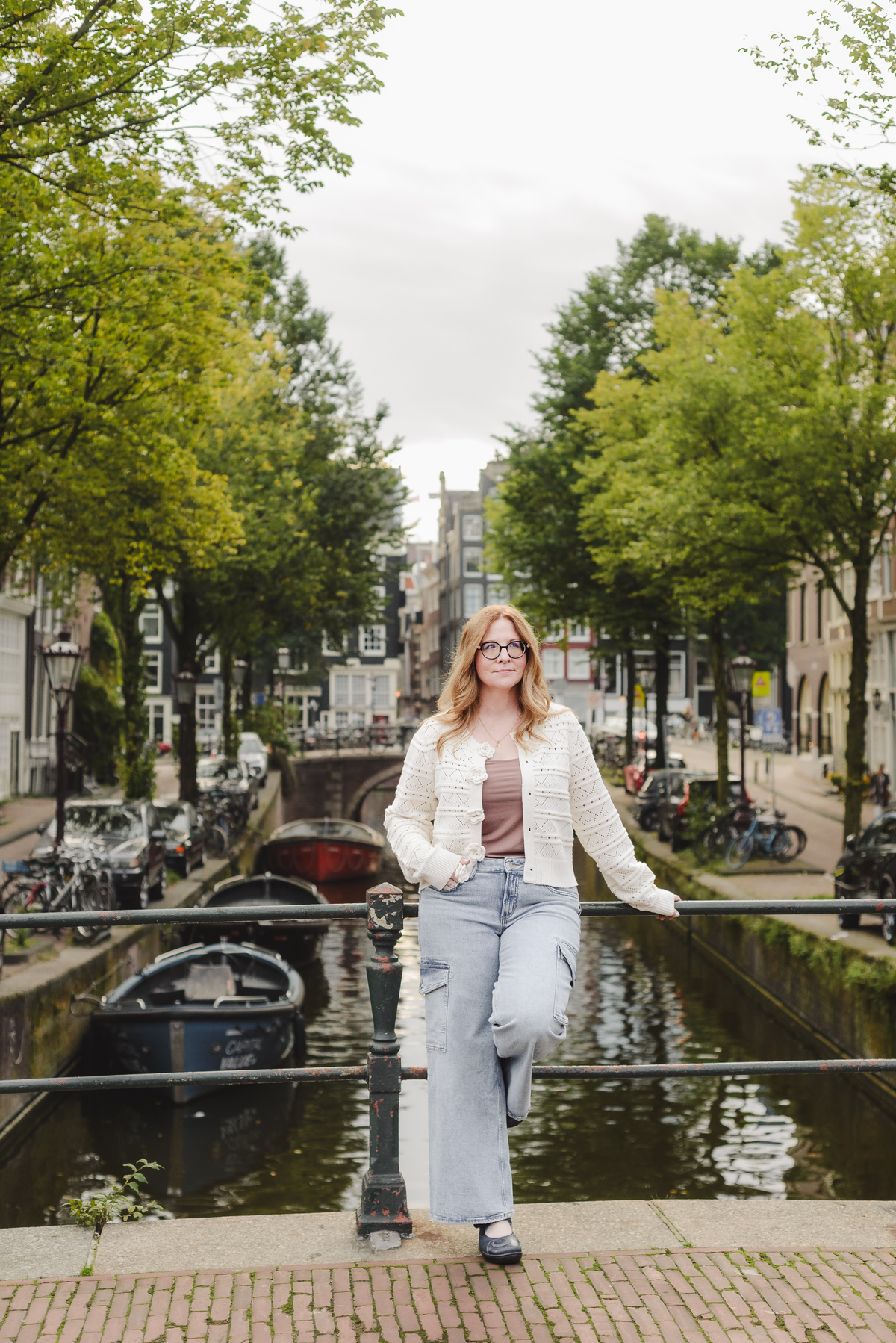 Female business owners capturing her trip in Amsterdam. She is in front of a canal with beautiful historical houses, trees aligned on each side, and boats in the background 