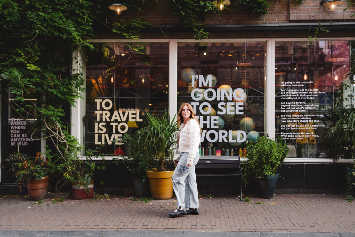 A female travel agent, capturing her trip in Amsterdam through a vacation photography session, is walking down the street in Amsterdam in front of a store whose messages are shown in the background: 