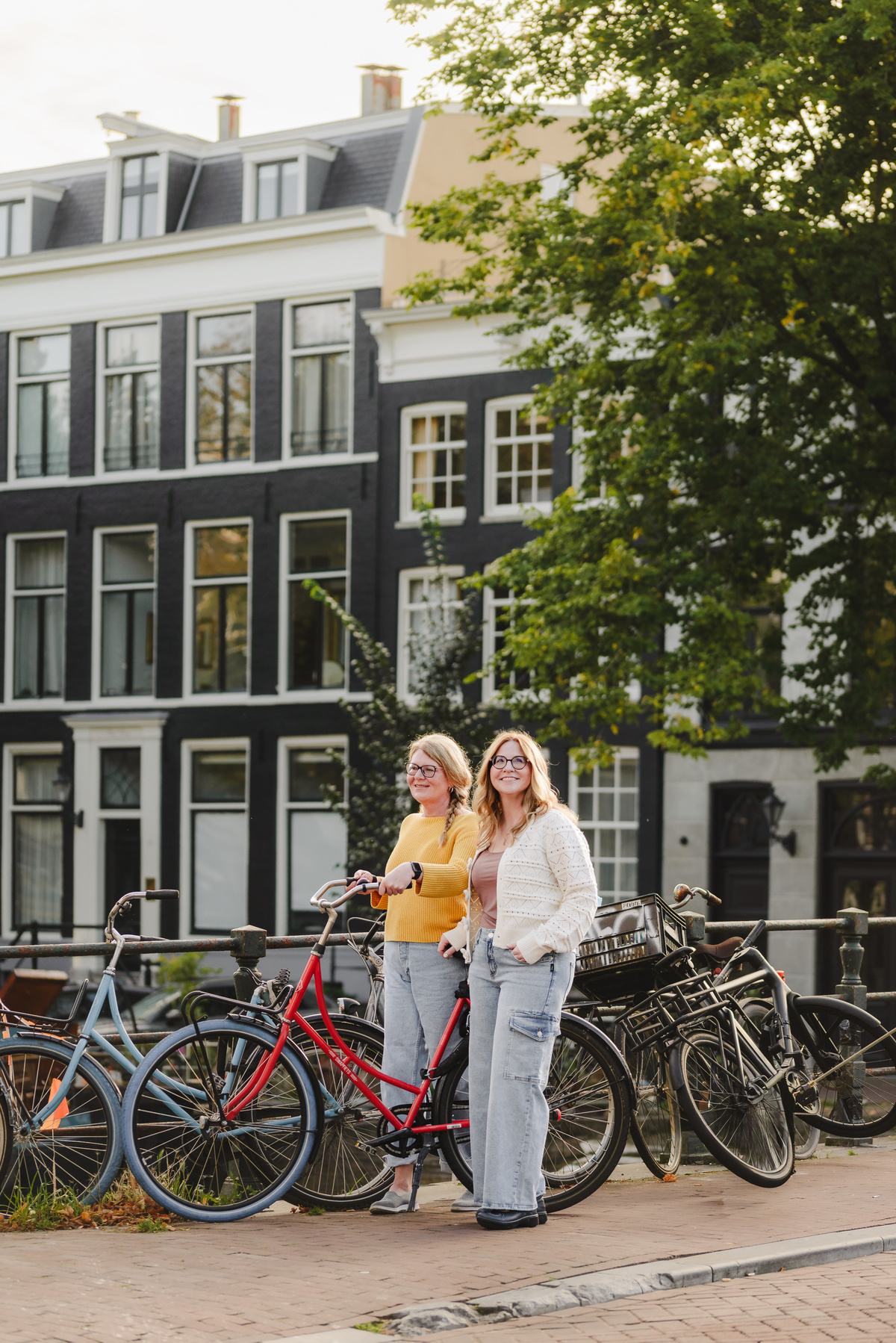 Sisters posing in front of a canal with historical houses and full of bikes in the background, with beautiful flowers too. They are holding red bike, and enjoying their time in Amsterdam. Vacation Photography session in Amsterdam, family photos.