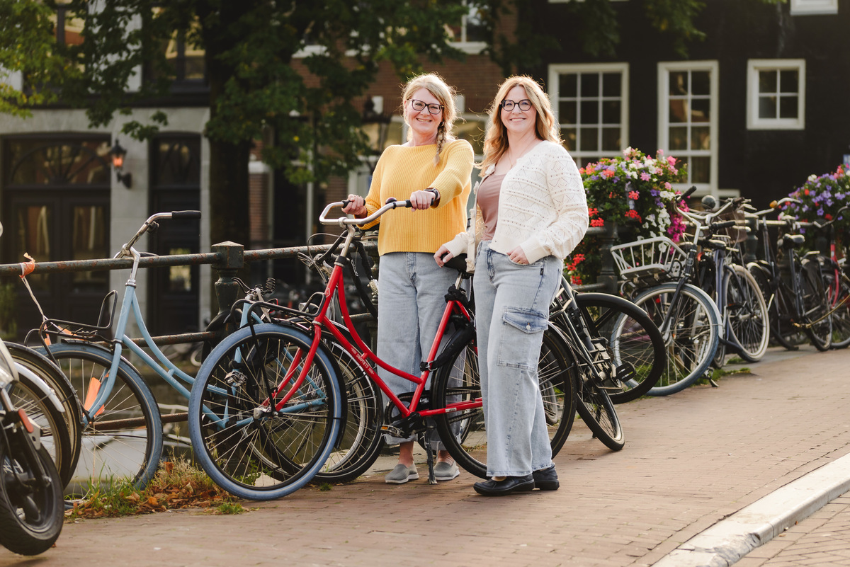 Two sisters holding a bike while posing for her photo, vacation photography photo session in a canal in Amsterdam. They are holding a red bike while smiling to the camera. 