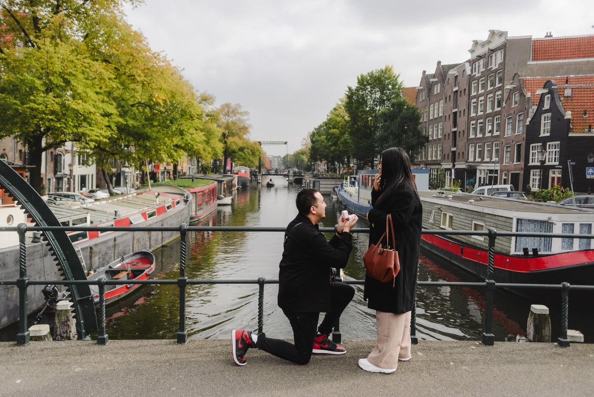 Man proposing beside an Amsterdam canal with historic houses and a traditional boat in the background.