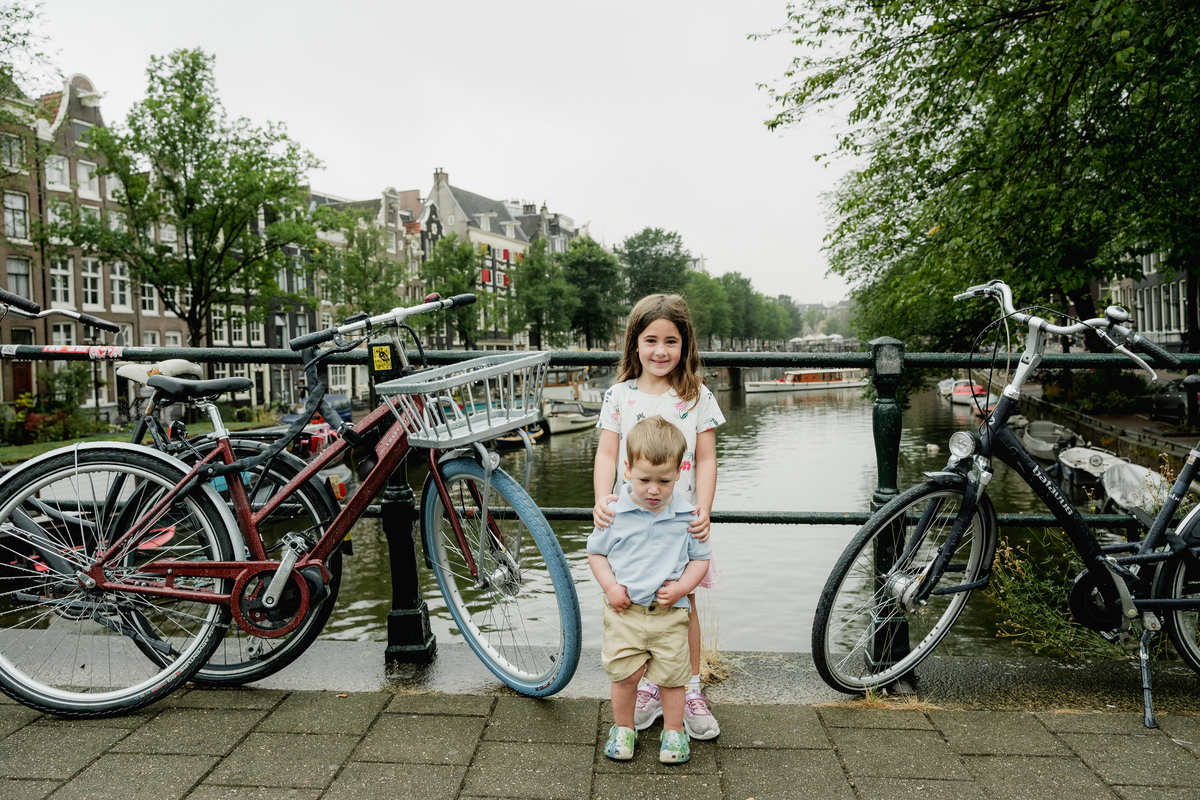 Siblings standing beside parked bicycles on an Amsterdam canal bridge.