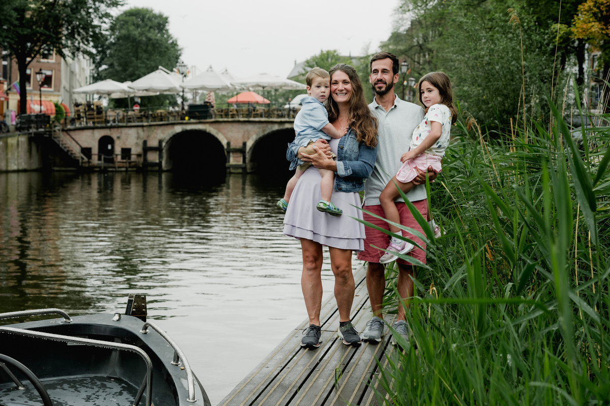 Parents holding their children beside an Amsterdam canal, with an arched bridge behind them.