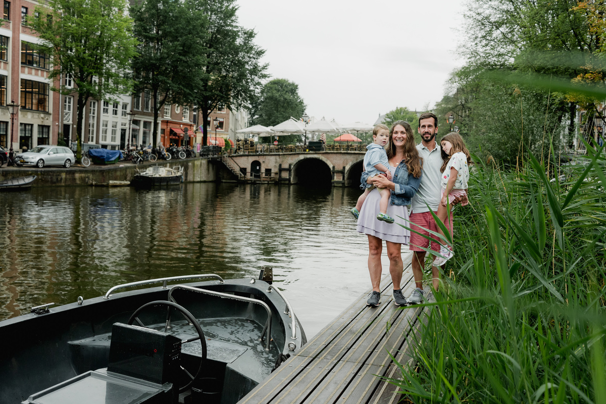 Wide view of a family beside the canal, framed by boats, greenery, and Amsterdam houses.