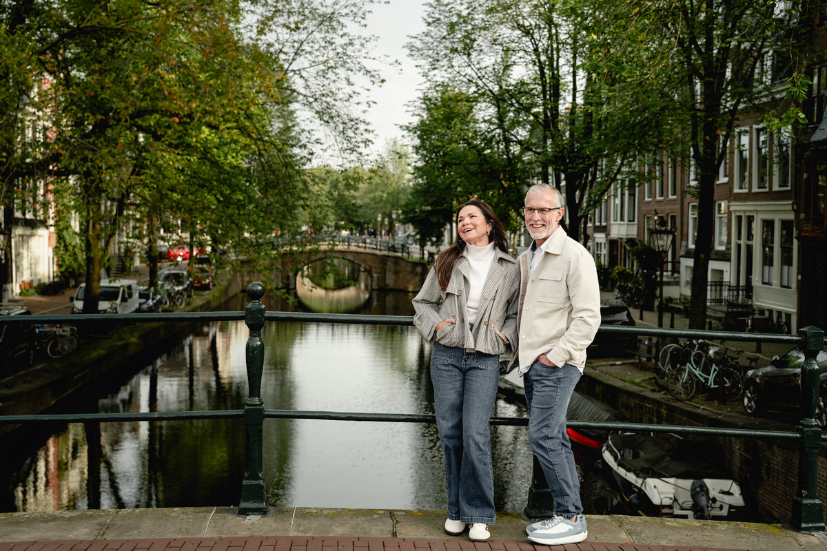 Couple standing on a bridge surrounded by trees and Amsterdam canal houses.