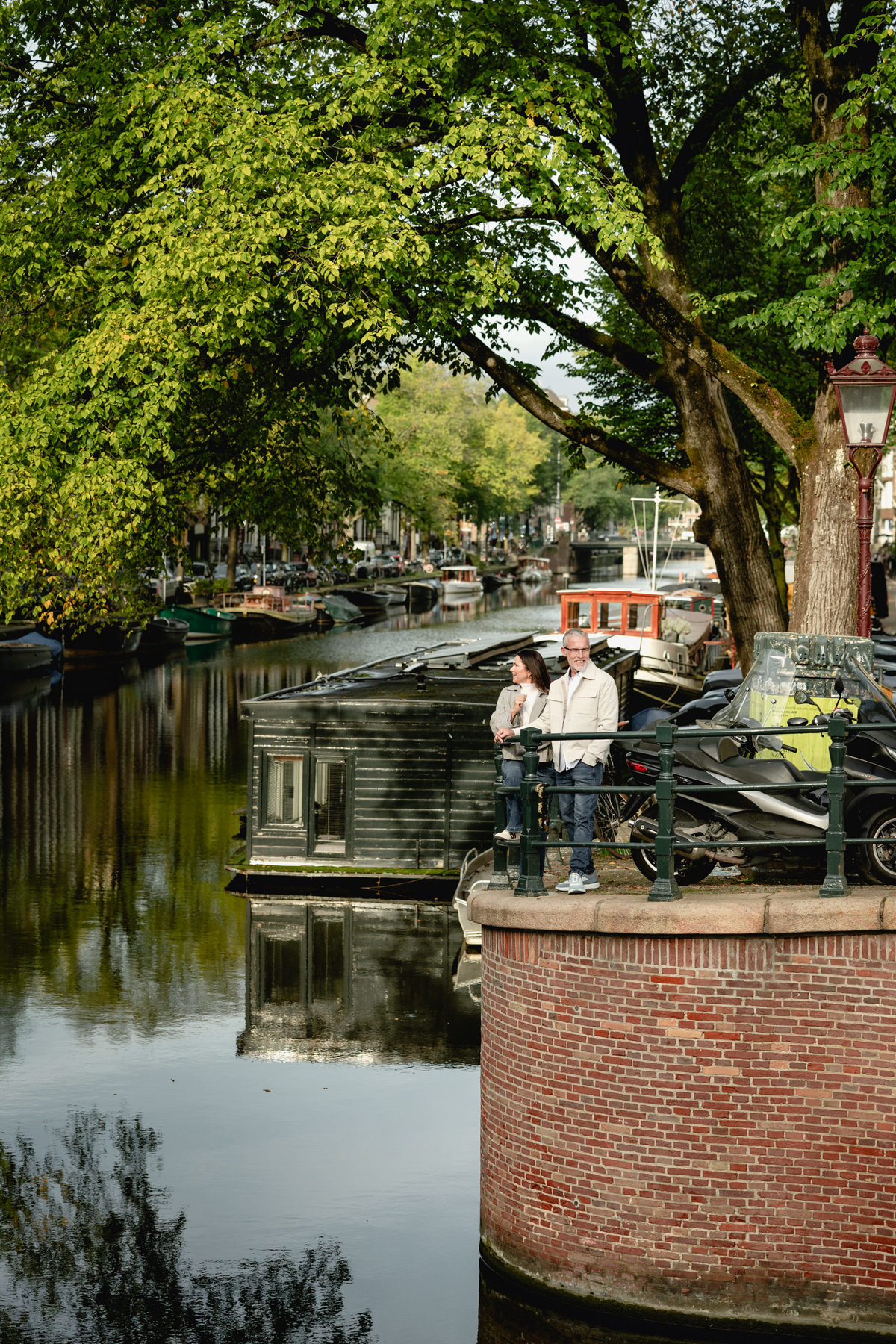 Couple beside a tree-lined canal with boats and historic houses behind them.