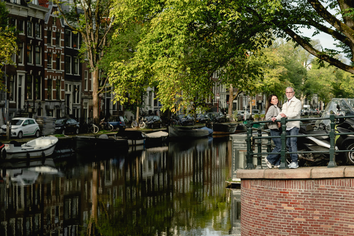 Couple standing together beside an Amsterdam canal, surrounded by green trees.
