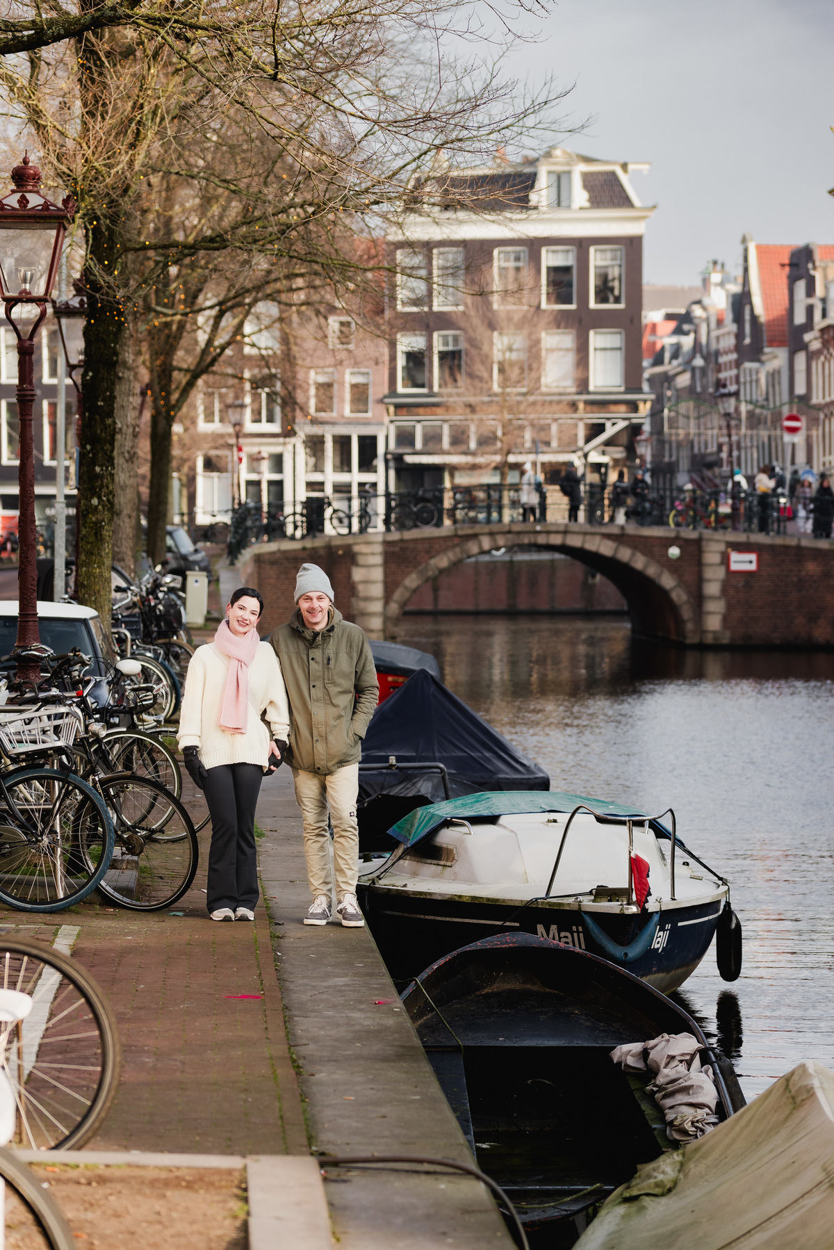 Couple standing beside an Amsterdam canal with historic houses and a boat behind them.
