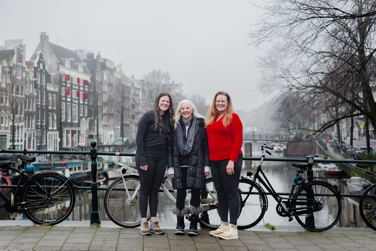 Three women standing together beside an Amsterdam canal with bicycles on a foggy winter morning.