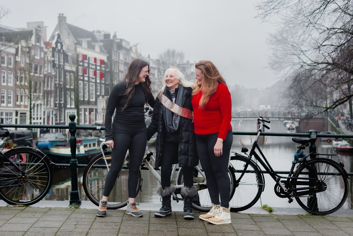 Three women smiling together beside an Amsterdam canal and bicycles