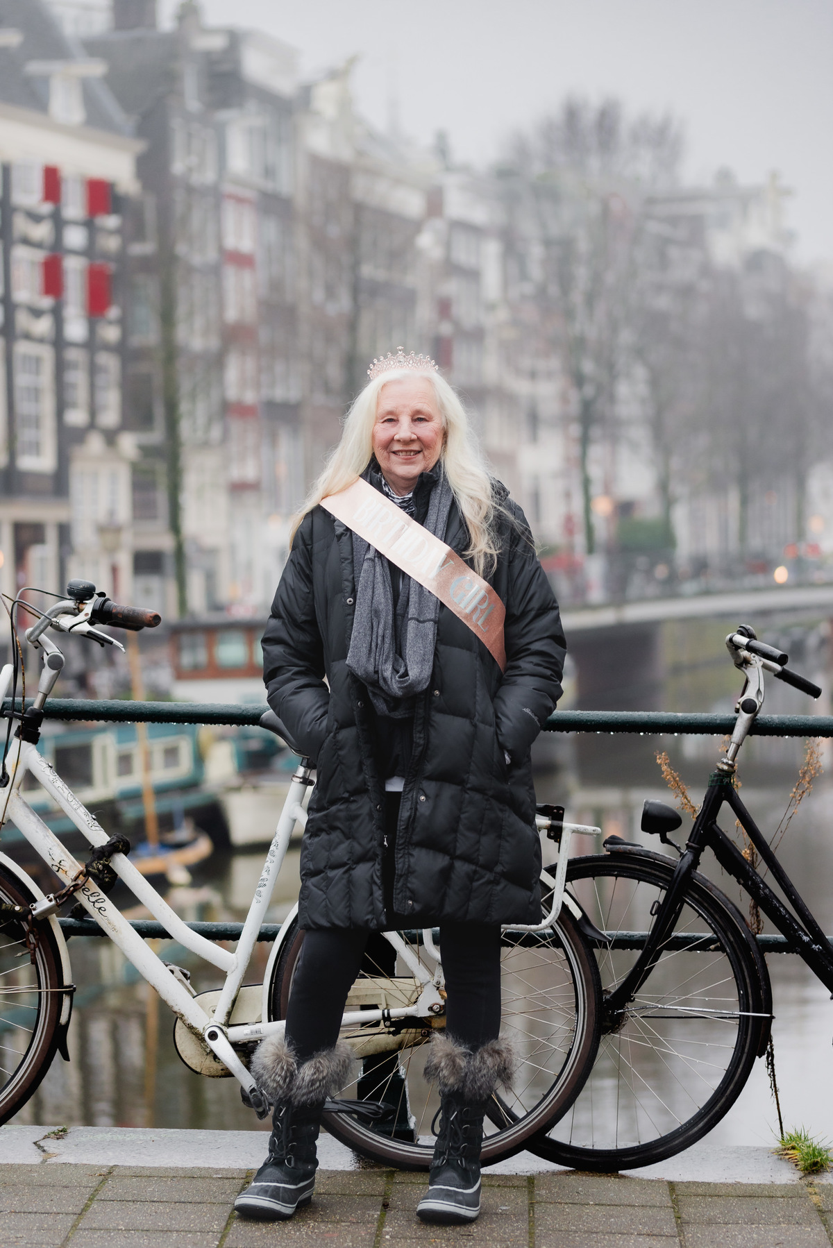 Woman in a birthday sash standing beside a bicycle on a canal bridge in Amsterdam.