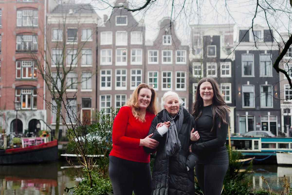 Three women standing together beside a canal with historic Amsterdam houses in the background.