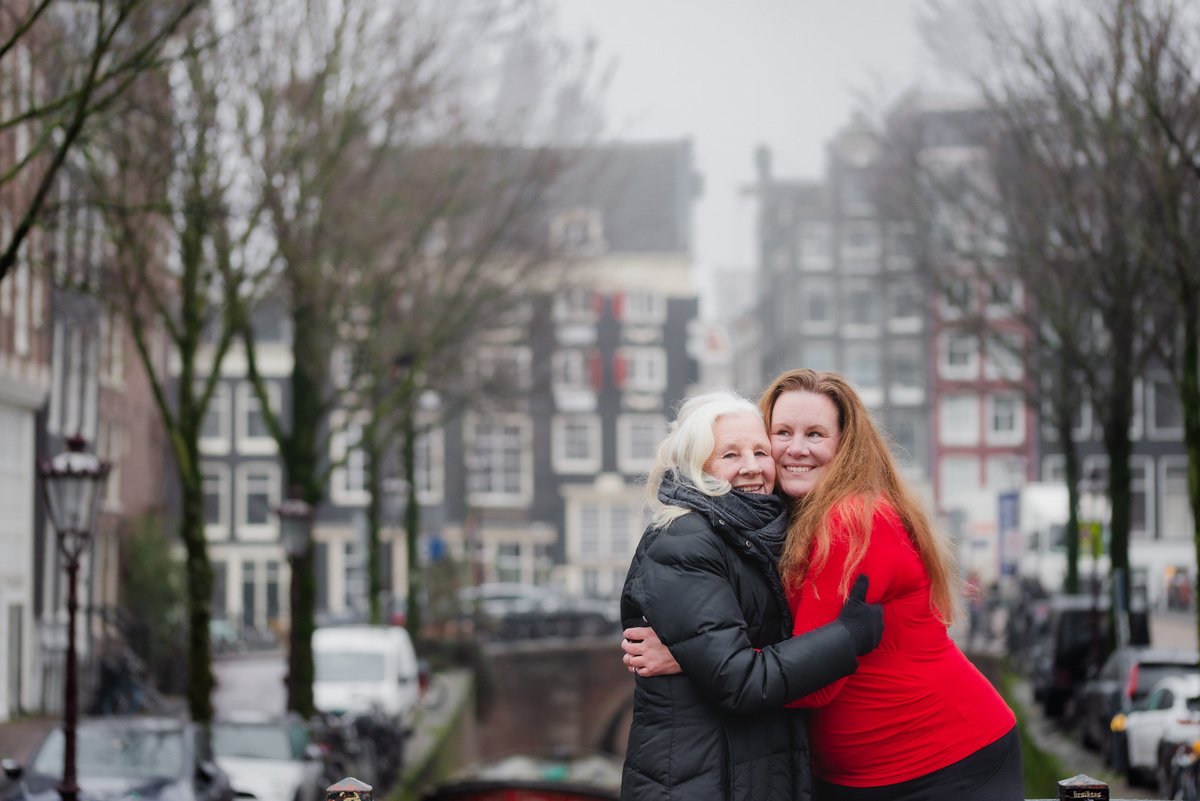 Two women hugging and smiling beside a quiet Amsterdam canal.