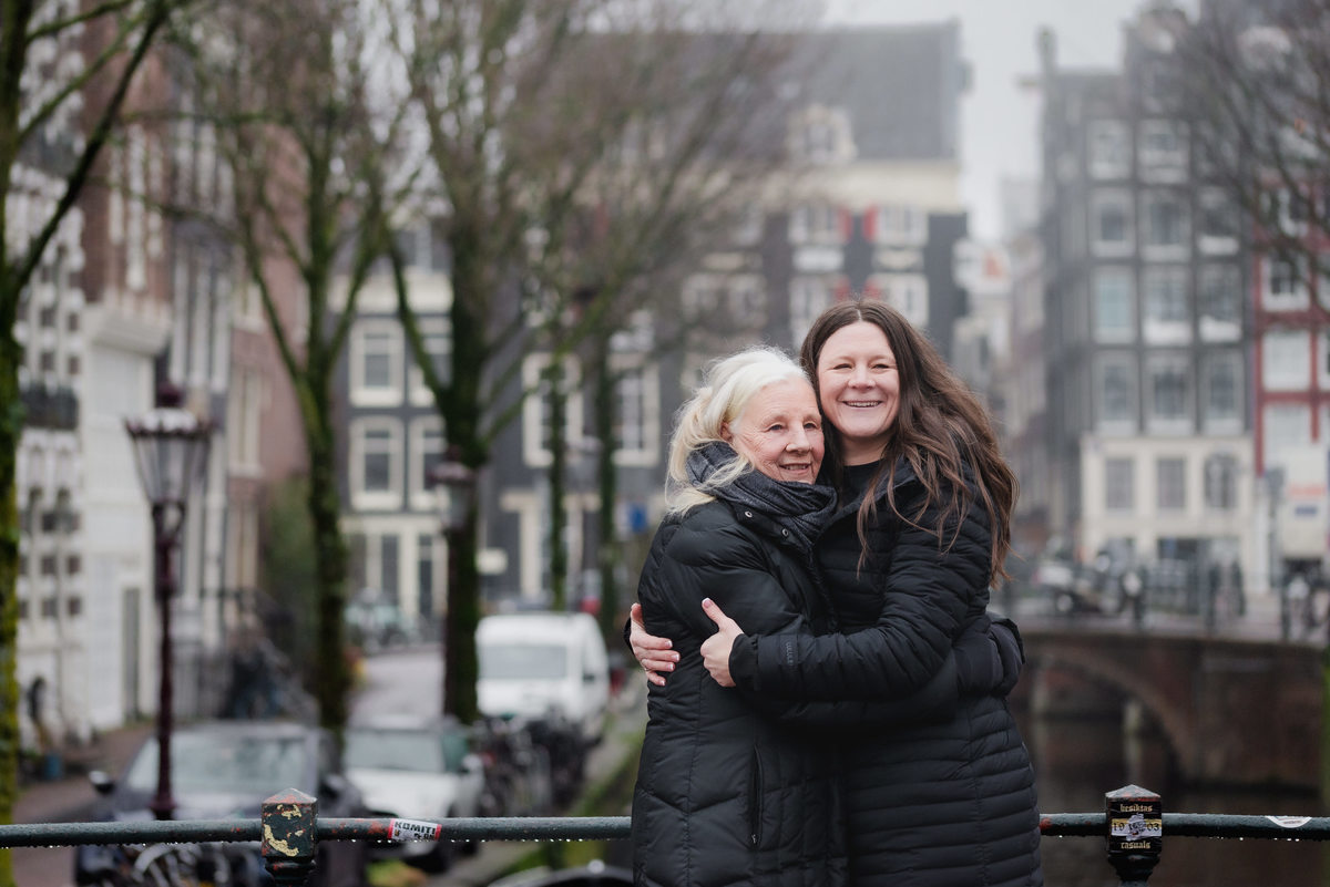 Two women embracing on a bridge with Amsterdam canal houses in the background.
