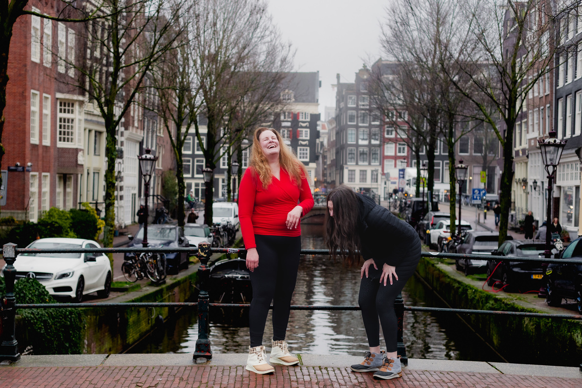 Woman in a red sweater laughing beside an Amsterdam canal with her sister during a family photo session.
