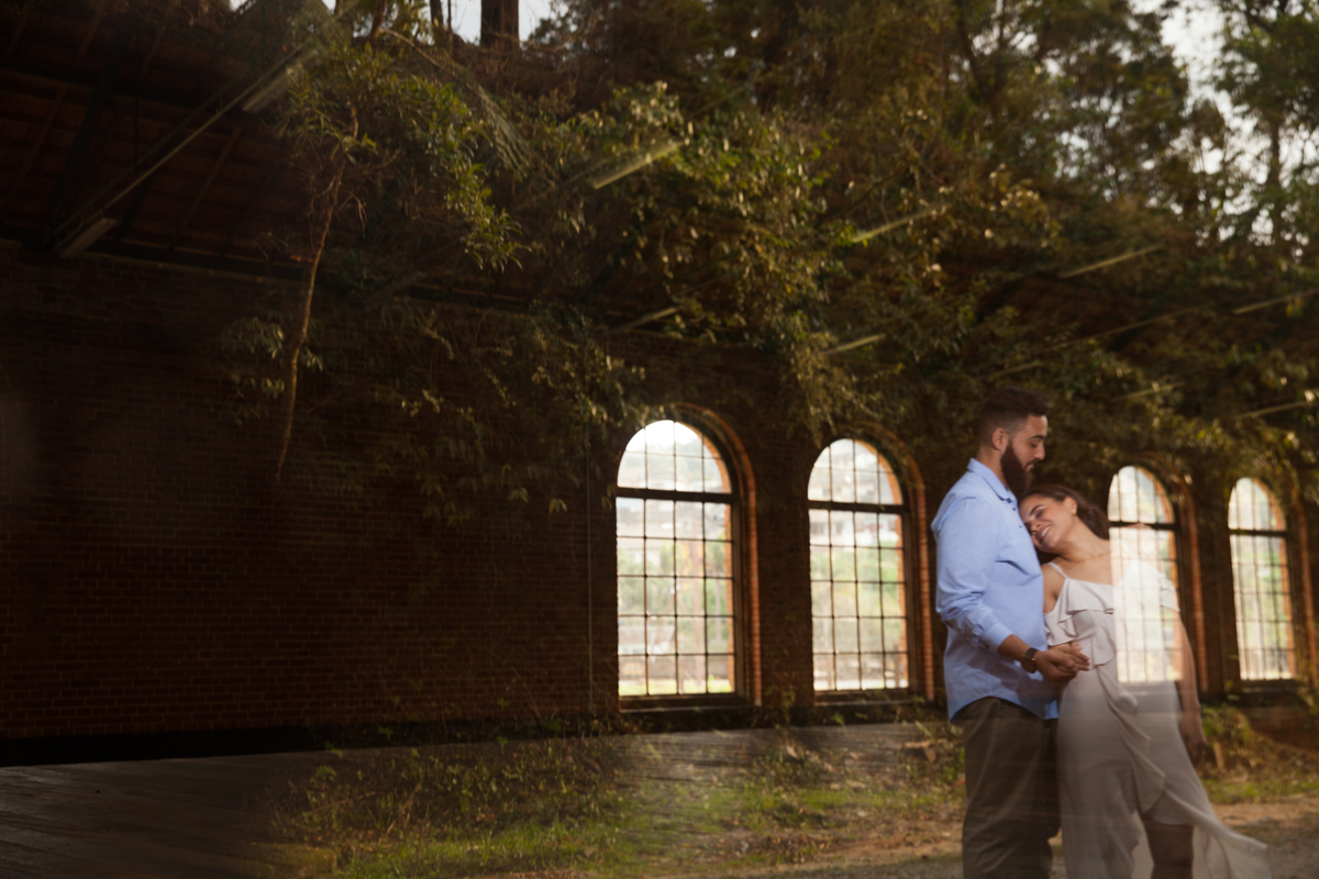 fotos de ensaio, pre wedding dos noivos em Paranapiacaba, noivo abracando a noiva, Paranapiacaba, São Paulo - Sp com fundo da parede de tijolinhos da antiga estacao de trem com reflexo no vidro