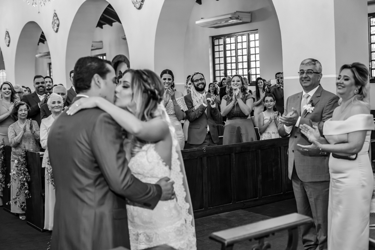 noivo beijando a noiva no altar e pais e convidados aplaudindo numa linda cena registrada pelo fotógrafo de casamento Campo Grande MS Vicente e Lilian Barros em foto preto e branco
